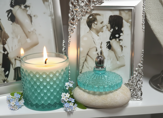 A small teal colored candle in a glass jar with a reflective lid, attached to a silver tassel, displayed on a shelf with two framed pictures in the background.