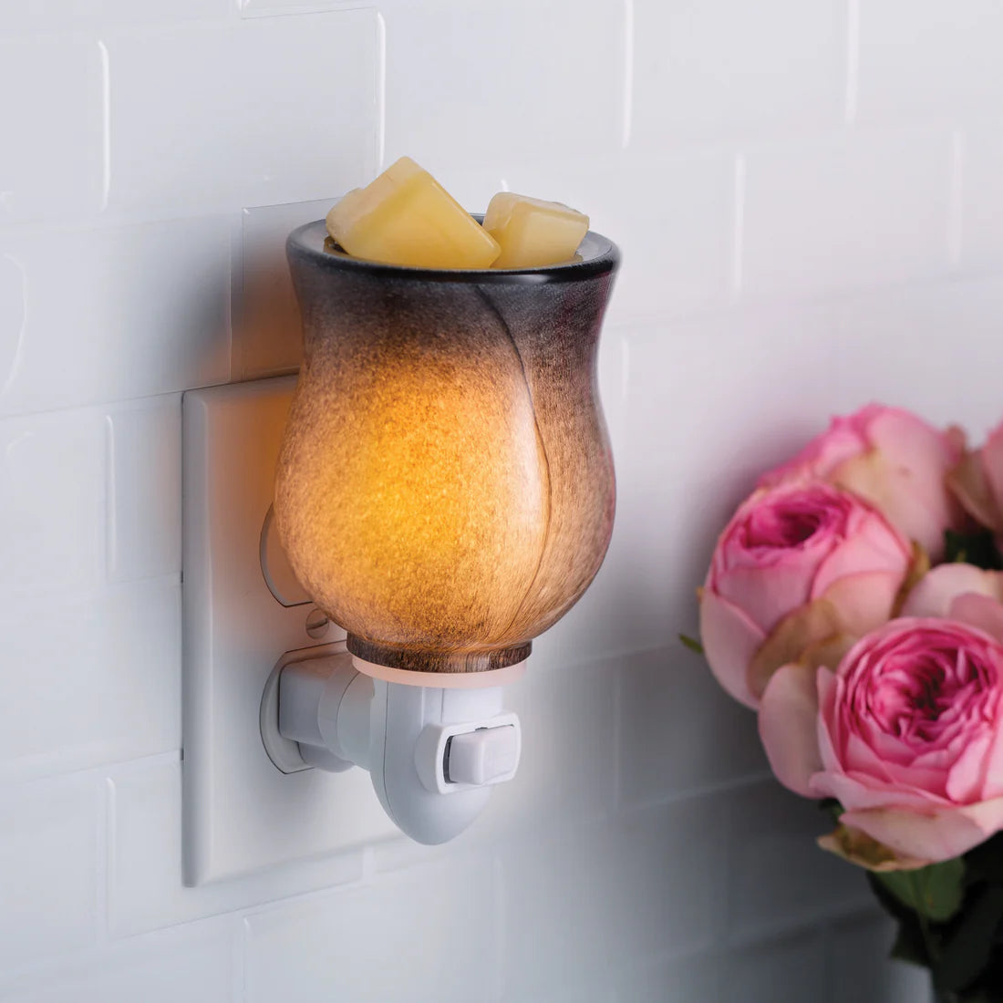 Wall-mounted wax warmer emitting a warm glow on a tiled wall with pink roses beside it.